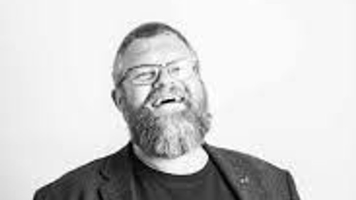 En skægget mand i jakkesæt og t-shirt med tryk poser med et glad udtryk mod en ensfarvet baggrund.