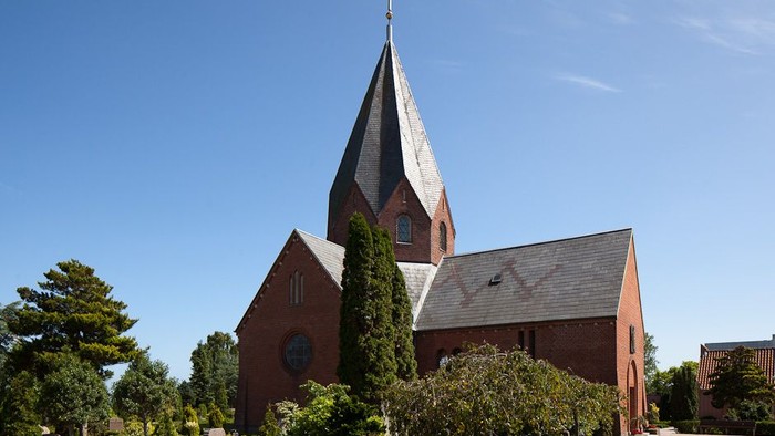 En rød kirke med spids tårn og kryds på toppen, omgivet af træer og buske under et klarblåt himmel.
