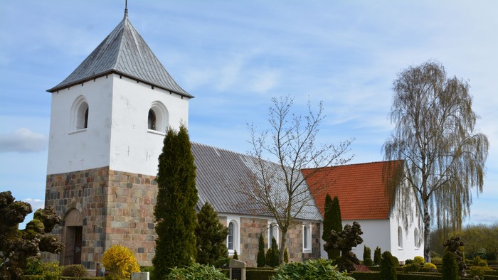 Hyggelig kirke med hvide mure, stensokkel og rødt tag omgivet af velplejet grønt og træer.