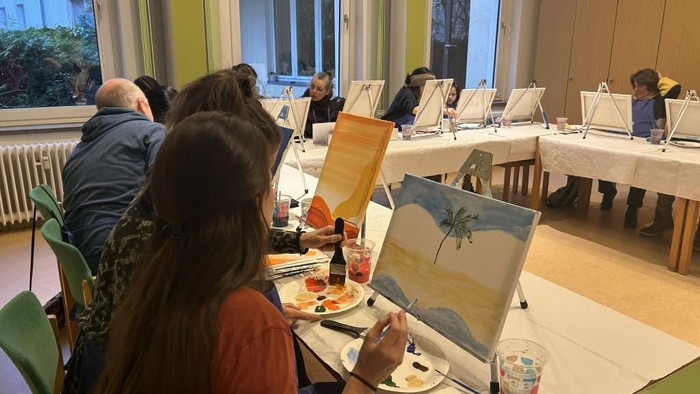 Ein Raum, in dem au fmehreren Tischen kleine Staffeleien stehen. Viele bunte Bilder und malende Menschen sind zu sehen.