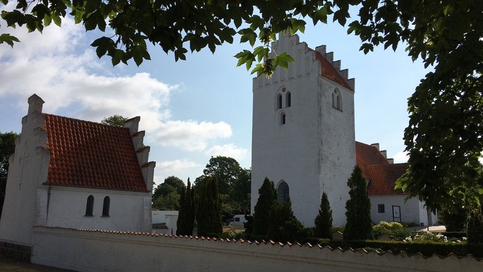 Et hvidt kirkebygningskompleks med røde tag og en tårn, omgivet af træer og en mur.