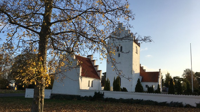 Et hvide kirke med spiret og tårn, omgivet af faldne løv og træer