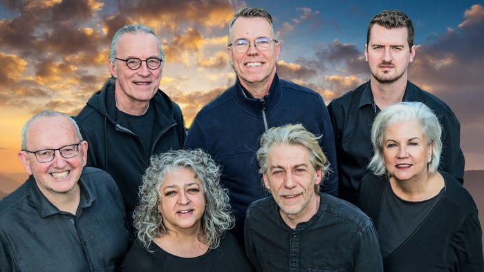 Eine Gruppe von sieben Personen posiert vor einem Himmel mit Wolken und Sonnenuntergang.