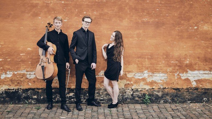 Tre musikere poser foran en rustik mur, hvor de holder og spiller på strengeinstrumenter i formelt tøj.