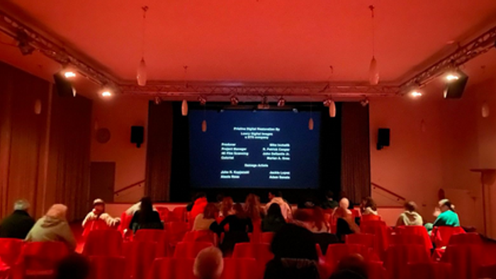 Ein vollbesetzter Hörsaal mit roten Sitzen, der eine Präsentation oder Vorführung auf einer großen Leinwand verfolgt.