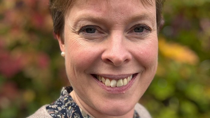 En glad kvinde smiler udendørs foran en farverig efterårsløvbaggrund.