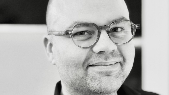 En smilende skaldet mand med briller iført en sort skjorte foran en moderne geometrisk baggrund.