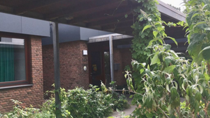 rotes Backsteinhaus mit Flachdach und überdachtem Weg zum Eingang, rechts und links davon begrünt