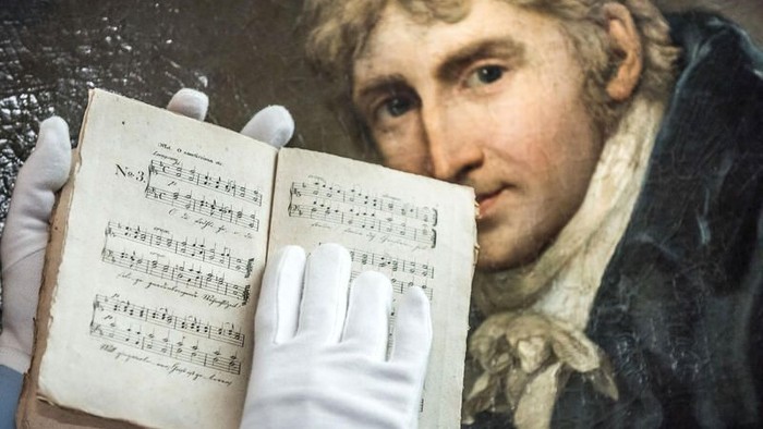 Eine behandschuhte Hand hält ein altes Notenbuch vor ein Porträt eines klassischen Komponisten.