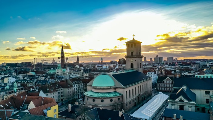 Et panoramisk bylandskab ved solnedgang med historisk arkitektur og en blanding af tage under dramatiske himmelstrøg.