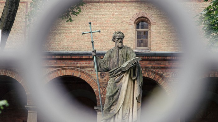 Statue einer bekleideten religiösen Figur, die einen Stab und ein Kreuz vor einem gewölbten Backsteingebäude hält.