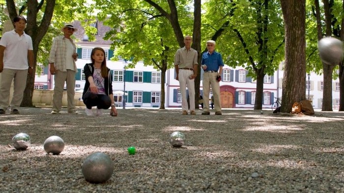 Eine Gruppe von Menschen spielt Boccia auf einem Kiesplatz unter Bäumen.