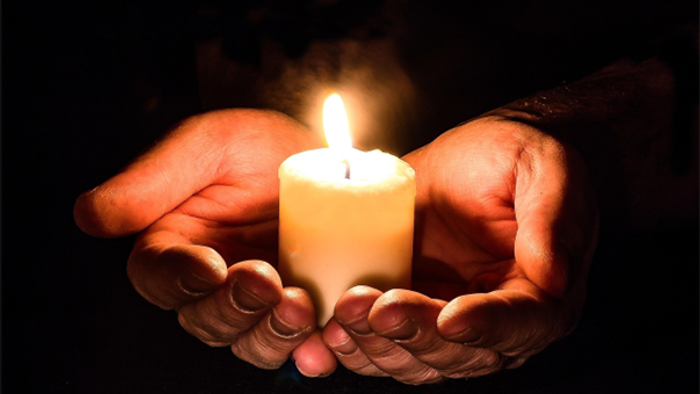 Hands gently cradle a lit candle in a darkened setting, casting a warm glow.