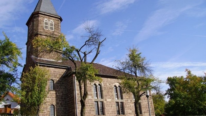 Kirche mit hohem Kirchturm und Rundbogenfenstern, umgeben von Bäumen und einem Metallzaun.