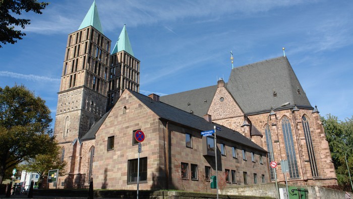 Große Kirche mit Zwillingstürmen und einem Kreuz obenauf, umgeben von Bäumen und Gebäuden.