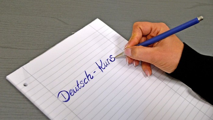 Eine Hand schreibt „Deutsch-Kurs“ auf einen linierten Notizblock mit einem blauen Stift.