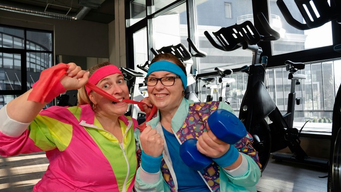 wei erwachsene Frauen sitzen eng nebeneinander in einem Fitnessstudio und blicken lachend in die Kamera. Beide tragen bunte Sportkleidung im Stil der 1980er-Jahre mit Stirnbändern und farblich auffälligen Trainingsjacken. Die Frau links hält ein rotes Fit