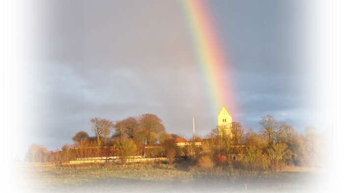 En levende regnbue buer sig over et malerisk landskab med et fjernt kirkespir og efterårsfarvede træer.