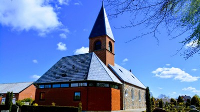 En rød kirke med spids tårn og skråt tag, omgivet af grønne buske og et klart blåt himmel.