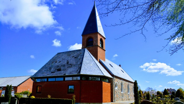 En rød kirke med spids tårn og skråt tag, omgivet af grønne buske og et klart blåt himmel.