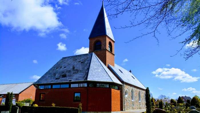 En rød kirke med spids tårn og skråt tag, omgivet af grønne buske og et klart blåt himmel.