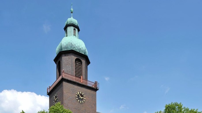 Historische Backsteinkirche mit grünem Kupferdach und Uhrturm, umgeben von üppigen grünen Bäumen.