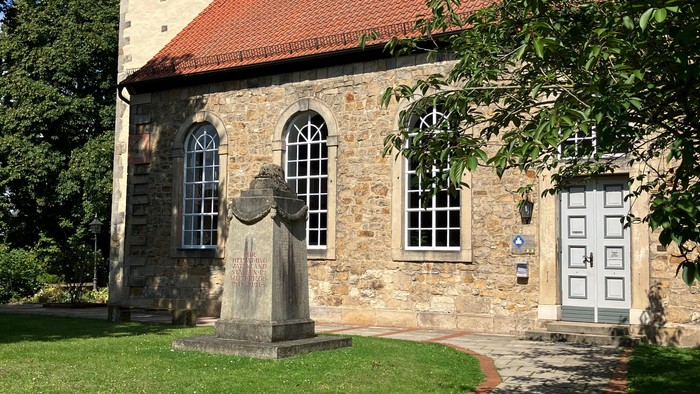 Kleine Kirche mit rotem Dach und Kreuz auf dem Turm, umgeben von Bäumen und Gras.