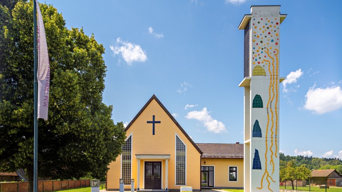 Gelbe Kirche mit hohem, buntem Turm und Kreuz an der Spitze, umgeben von Bäumen und Gras.
