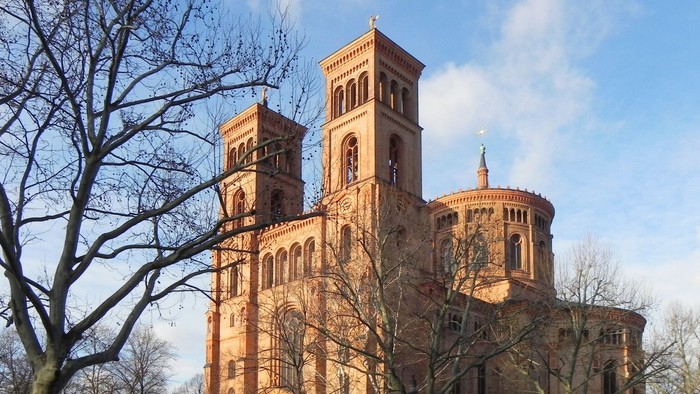 Großes, altes Backsteingebäude mit zwei Türmen und Rundbau, umgeben von kahlen Bäumen und einem Auto