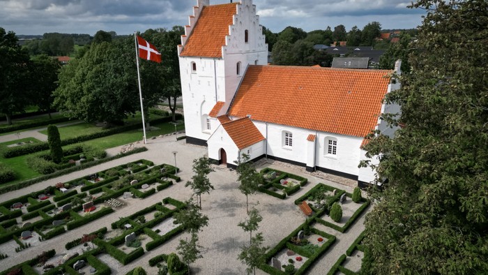 En hvid kastedal med røde tag og en flagstang med danskirkeflag i en park med formede beder og grønne arealer