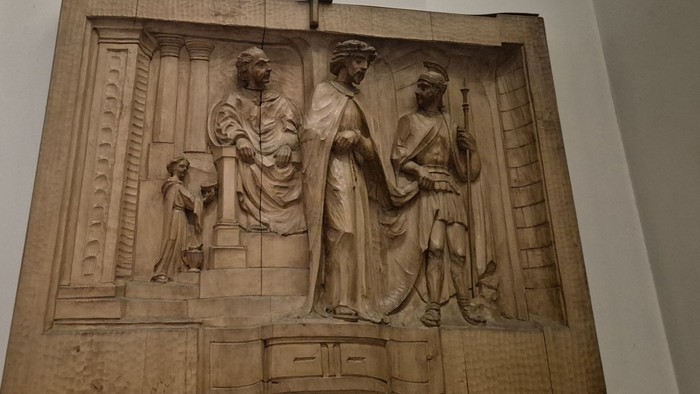 Reliefskulptur, die drei klassische Figuren im Dialog mit architektonischen und symbolischen Elementen darstellt.