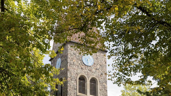 Der Kirchturm aus Stein mit Uhr und Fenster steht in einem Park.
