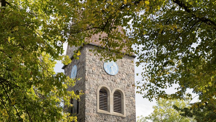 Der Kirchturm aus Stein mit Uhr und Fenster steht in einem Park.