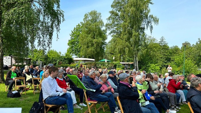 Eine Gruppe von Menschen sitzt im Freien auf Stühlen und hört einem Redner zu.