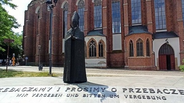 Gedenkstatue mit zweisprachiger Bitte um Vergebung vor einem historischen Backsteinkirchengeb�ude.