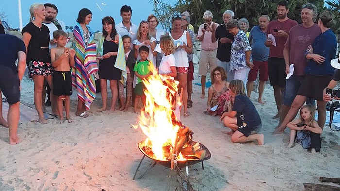 En gruppe af mennesker og børn samlet om et bål til Sankthans på en strand.