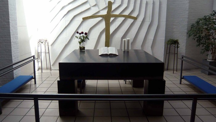 En slem med blomster og kande foran et stort kors på en muret vegg