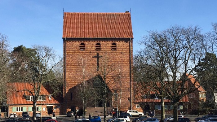 Ein rotes Backsteingebäude mit einem Kirchturm und einem Kreuz, umgeben von Bäumen und Autos.