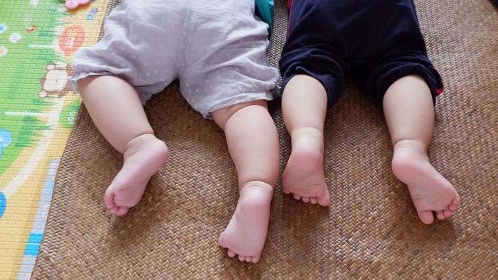 Zwei Babys liegen auf einer Matte, tragen Windeln und kurze Hosen, wobei Füße und Beine sichtbar sind.