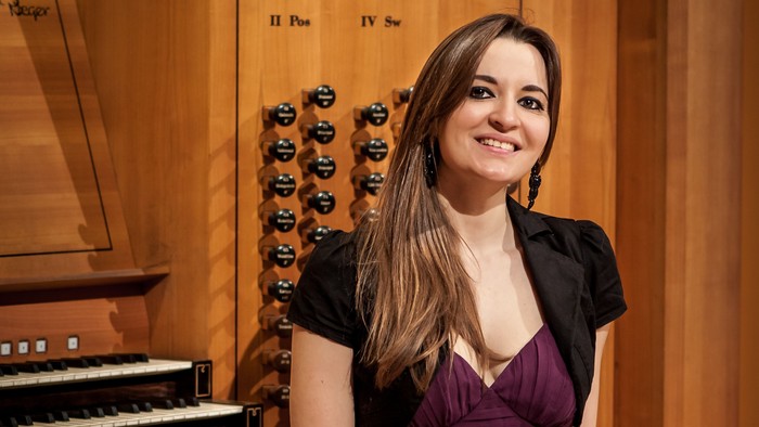 Eine Frau in einem violetten Kleid sitzt lächelnd neben einer großen, verzierten Pfeifenorgel.