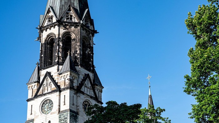Ein hoher Kirchturm mit Uhr und spitzen Bögen vor blauem Himmel und Bäumen.