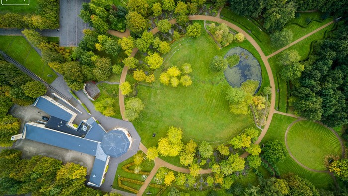 En stor park med en rund sø og en stor blomsterbede i midten, omgivet af træer og en bygning til venstre.