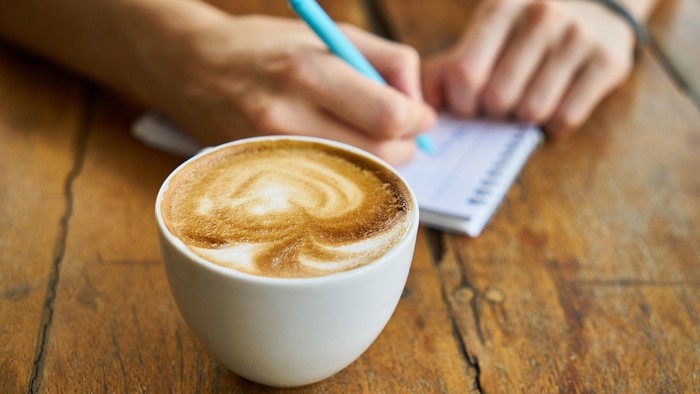 Eine Person schreibt in ein Notizbuch, während sie einen Latte-Art-Kaffeebecher mit einem Stift in der Hand hält.