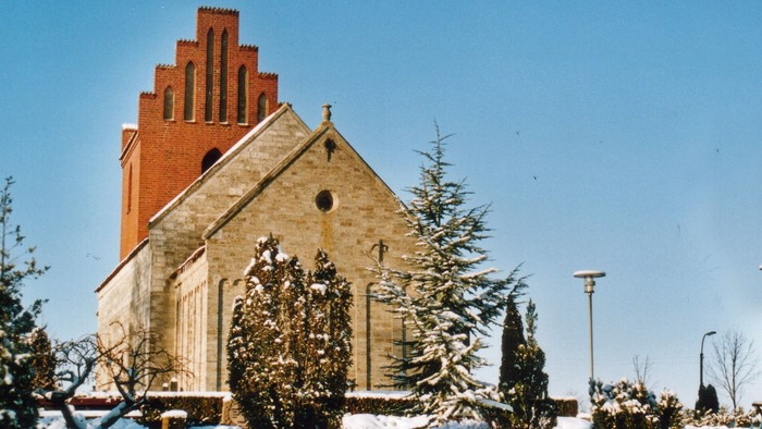 En snedækket kirke med teglmur og tårn, omgivet af snebede træer og en klar blå himmel