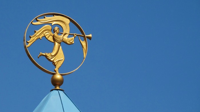 Goldener Wetterhahn mit einer engelhaften Figur, die eine Trompete hält, auf einem blau gedeckten Gebäude.