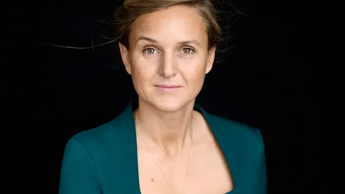 En kvinde i en turkis blazer poser med et neutralt udtryk mod en mørk baggrund.