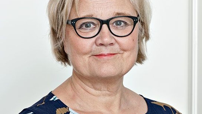 En kvinde med kort, blond hår og briller, smiler til kameraet.