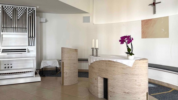 Moderne Kapelle mit Innenraum, der einen steinernen Altar mit Blumen, Kerzen und einer danebenstehenden Pfeifenorgel zeigt.