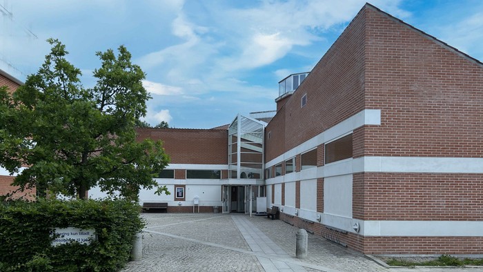 Et moderne bygning med røde mursten og hvide detaljer, beliggende ved en stenlagt gård med træer og buske.