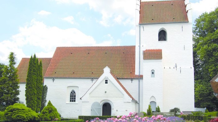 Et hvidt kirkebygning med røde tag og en tårn, omgivet af grønne planter og blomster.